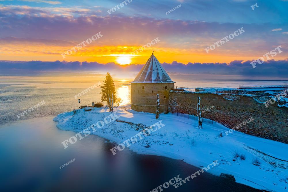 На фотографии изображена крепость Орешек, расположенная на острове в устье реки Невы. Зимний свет и краски рассвета подчеркивают древние каменные стены, добавляя теплоту и уют холодному пейзажу. Крепость Орешек, имеющая богатую историю, является одним из символов обороны северо-западных рубежей России.